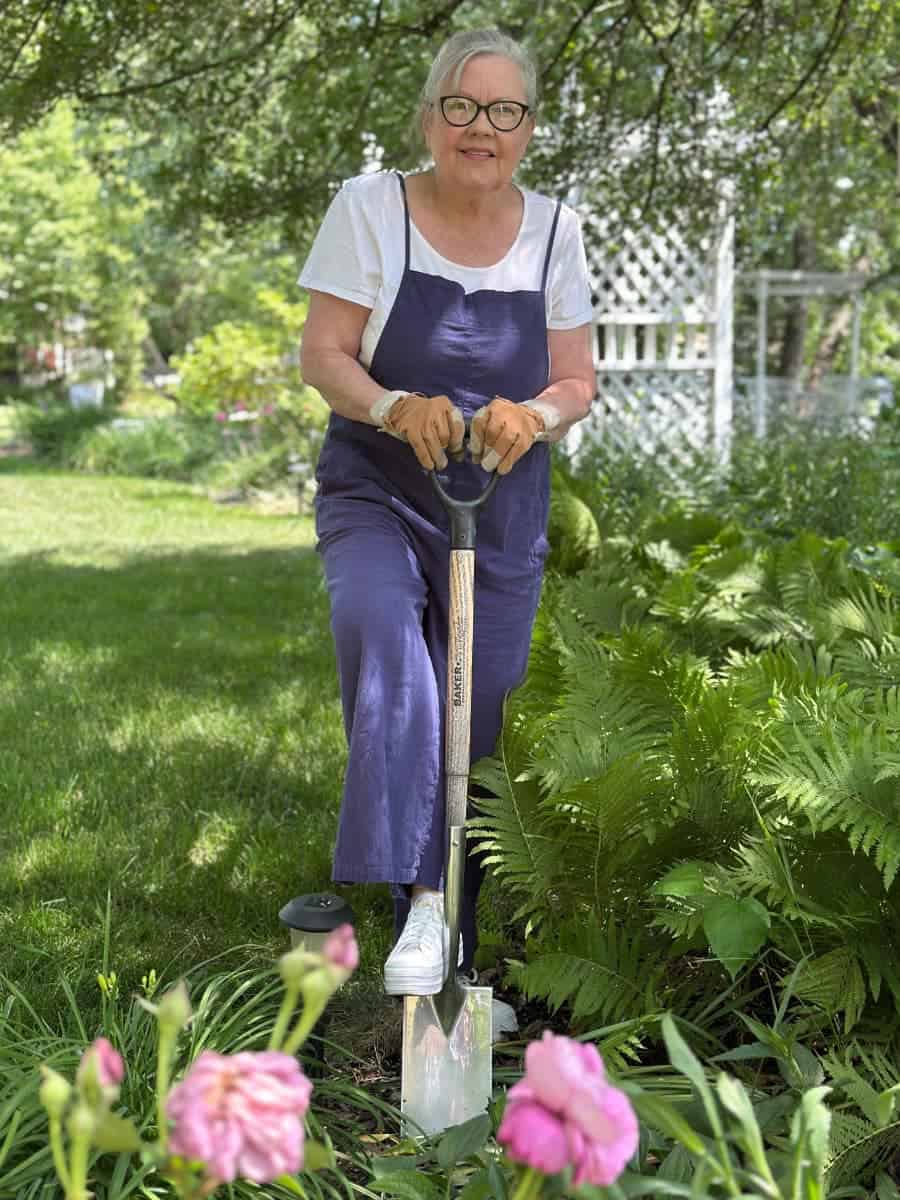 An older woman in a summer capsule wardrobe—white shirt, blue overalls, and gardening gloves—stands on grass, digging with a shovel among lush green plants and pink flowers. Trees and a white trellis frame the vibrant outdoor scene.