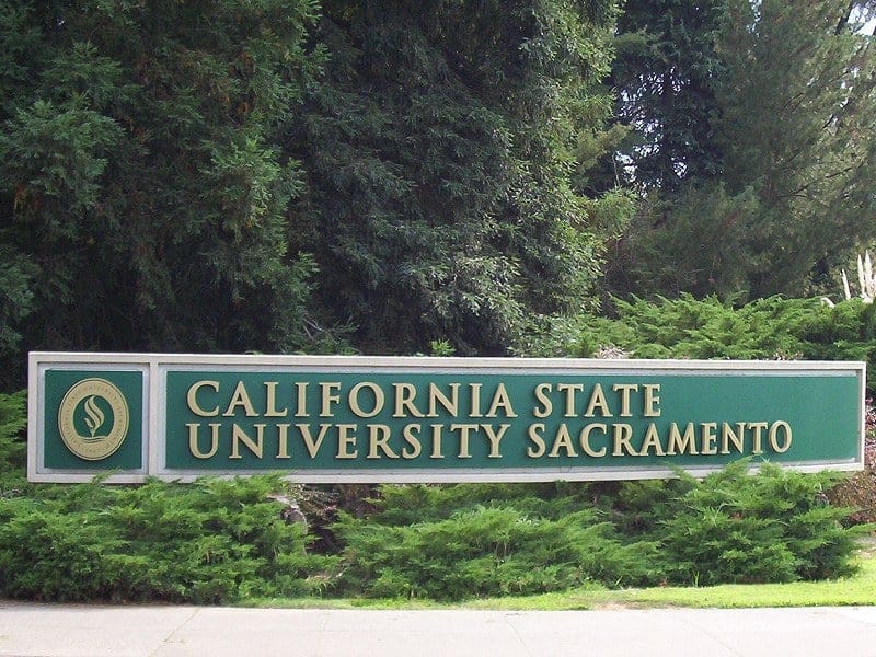 A green sign reading California State University Sacramento set in a low brick wall at a campus entrance, with trees behind it.