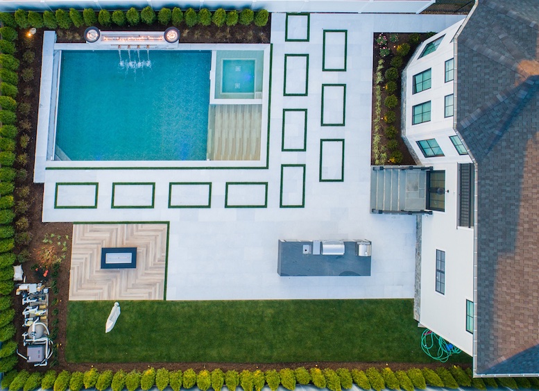 Aerial view of a modern backyard with a pool, hot tub, patio, outdoor kitchen, fire pit area, and landscaped green lawn.