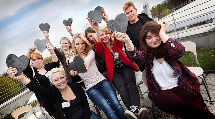 Mehrere junge Menschen sitzen zusammen und halten herzförmige Schiefertafeln in die Kamera, während sie lächeln und fröhlich posieren.