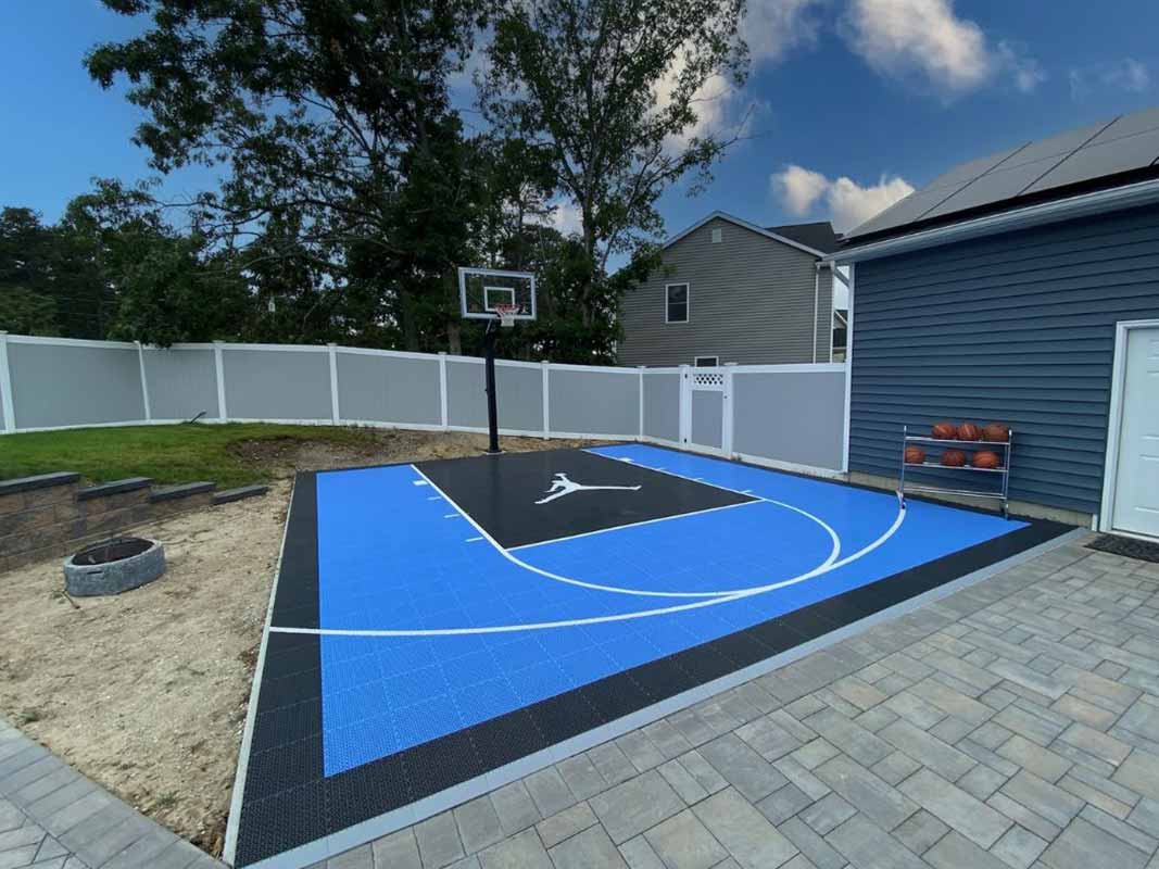 A backyard sports court installation in Huntington showcases a blue and black basketball surface, complete with a hoop and a rack of basketballs nearby. A white fence and trees create a picturesque backdrop.
