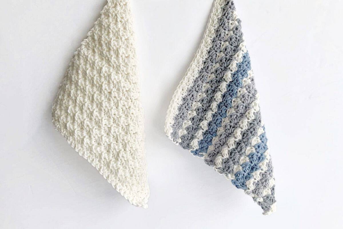 Two crocheted dishcloths with a bumpy, textured pattern hang on a white wall; one is solid off-white, and the other features stripes of white, grey, and blue. No visible text or promotional information appears in the image. Free crochet pattern
