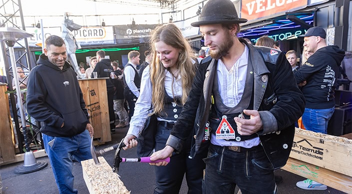 Zwei Menschen in historischer Kleidung stehen zusammen, einer hält einen pinkfarbenen Gegenstand, beide wirken interessiert und lächeln, im Hintergrund sind weitere Personen.