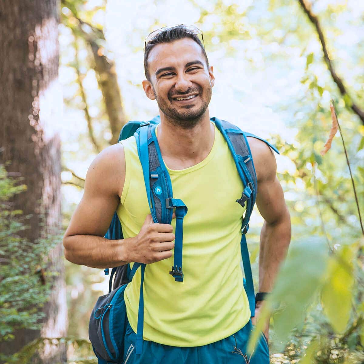 A man in a yellow tank top and blue backpack smiles while hiking in a sunlit, green forest.
