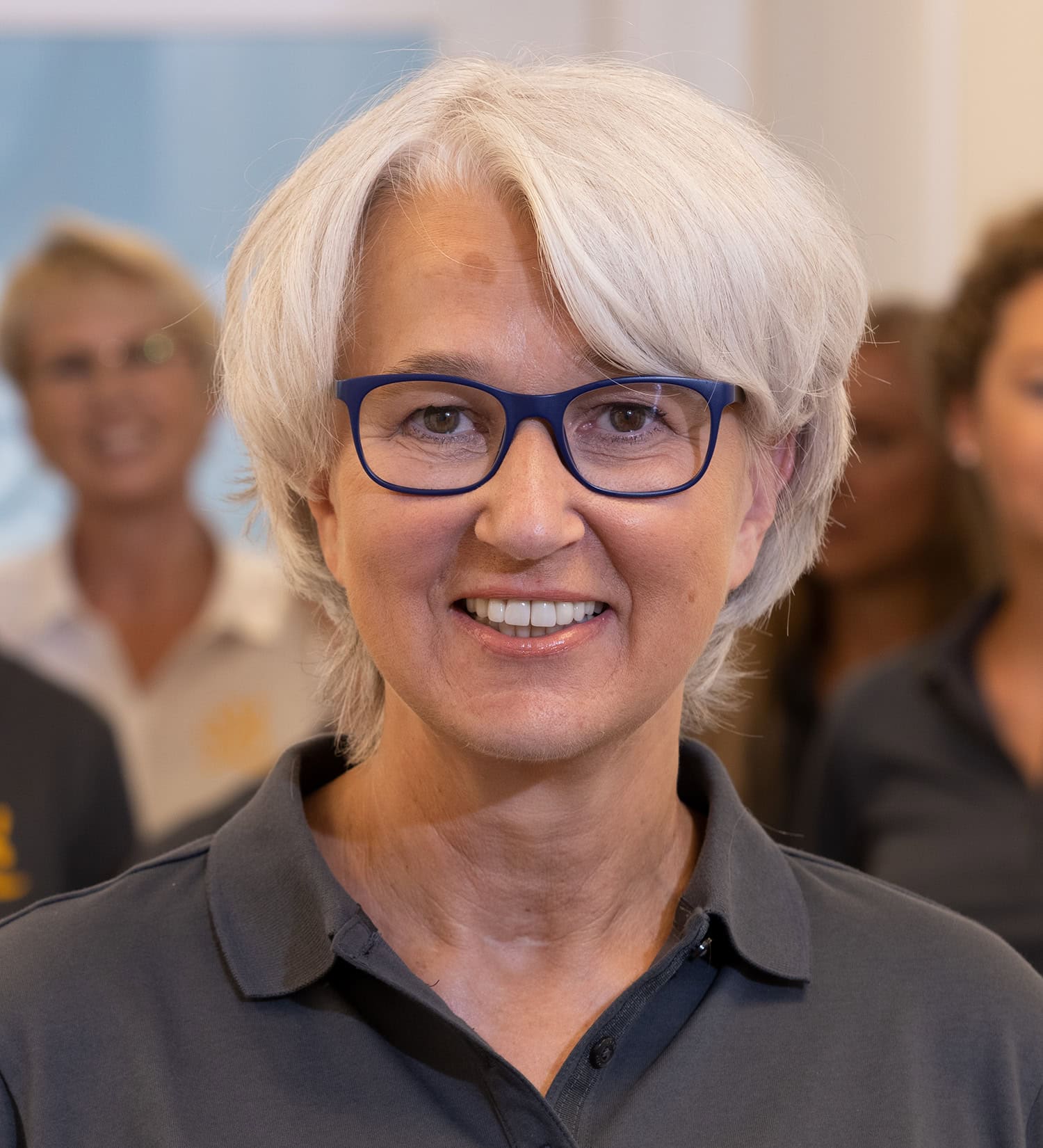 Lächelnde weisshaarige Mitarbeiterin mit Brille im Vordergrund vor dem Praxis-Team im Hintergrund.