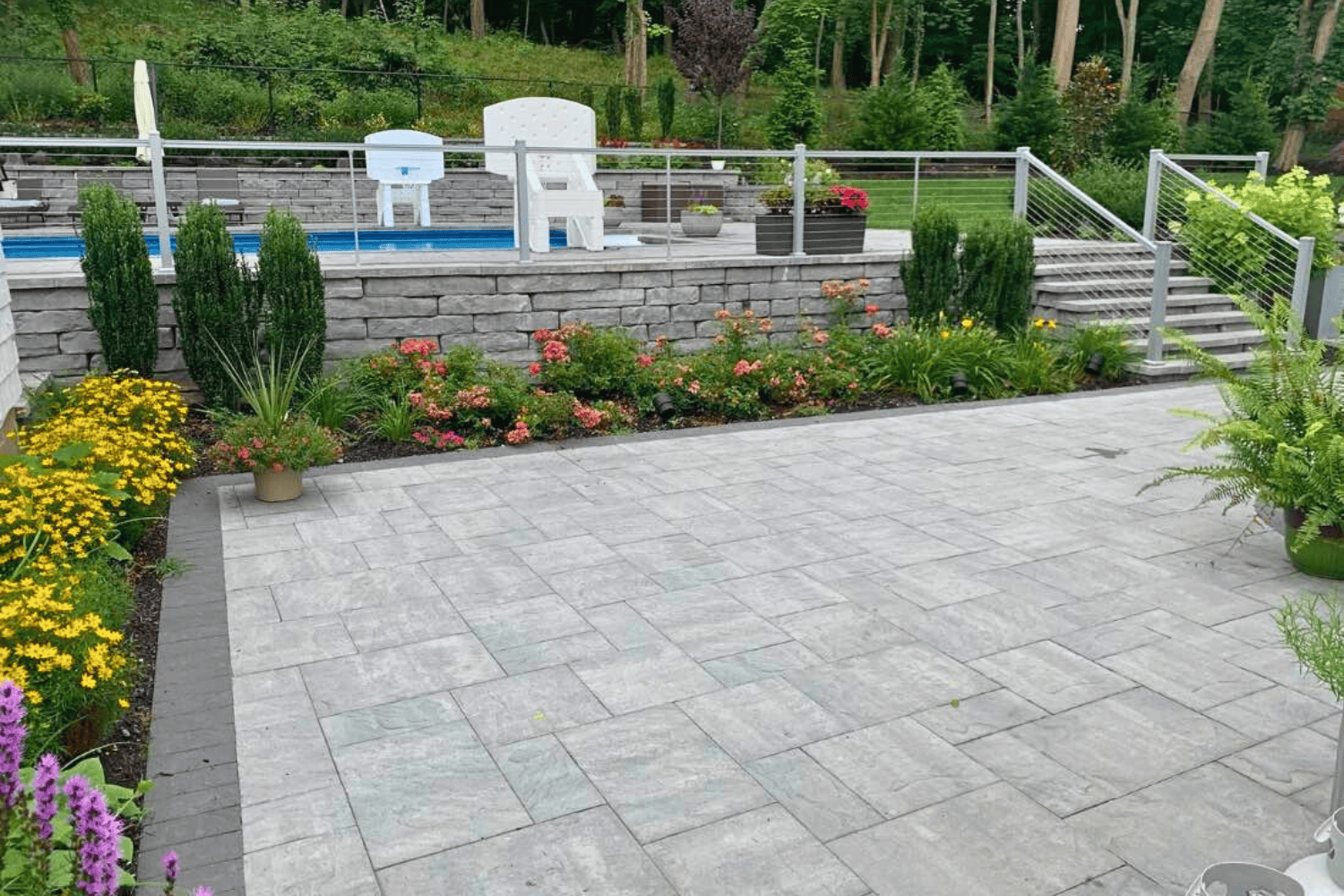 A landscaped backyard features a stone patio, colorful flower beds, and steps bordered by retaining walls, leading up to a pool area with chairs and a fence.