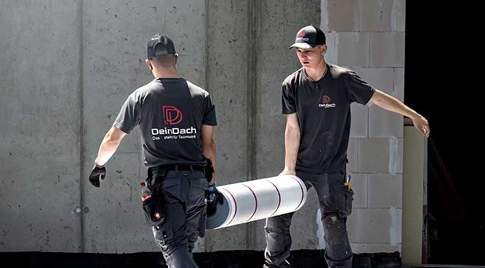 Zwei Arbeiter tragen zusammen eine große weiße Röhre vor einer Betonwand.