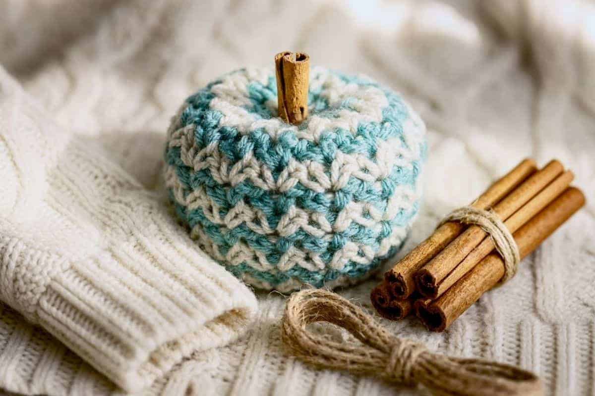 A blue and white yarn crochet pumpkin with a cinnamon stick stem sits next to bundled cinnamon sticks and a white textured sweater on a soft, cozy surface.