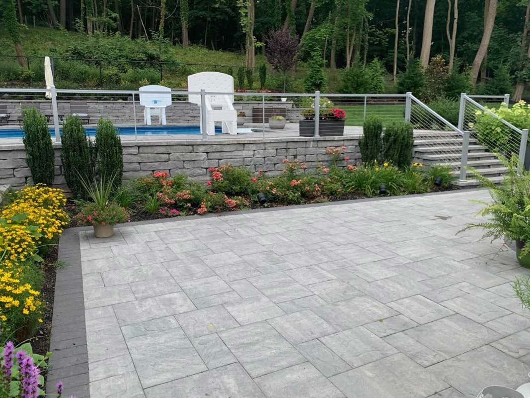 A backyard with a paved patio area and retaining wall, bordered by a garden with flowers and shrubs. Stone steps lead to an elevated area with a lounge chair and railings, and a pool is visible in the background in Elwood.