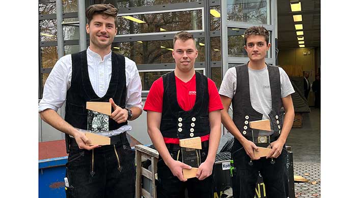 Ein junger Mann in rotem T-Shirt und traditioneller Zimmermannsweste hält ein Stück Holz, steht draußen vor einem modernen Gebäude.