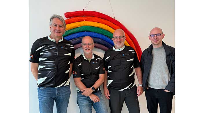Zwei Männer in schwarzen Radtrikots stehen vor einem Wandbild mit Regenbogenfarben.