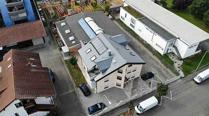 Bild von Firmensitz von Karl-Heinz Krawczyk eG Modernes Mehrfamilienhaus mit grauem Dach, Solarpanels und mehreren Fenstern, umgeben von anderen Gebäuden und parkenden Autos, aus der Vogelperspektive fotografiert.
