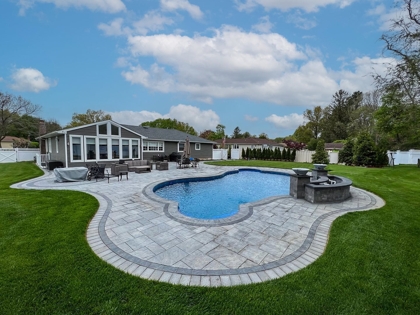 A backyard with a stone patio, in-ground swimming pool, spa, lawn, patio furniture, and a single-story house in the background on a partly cloudy day.