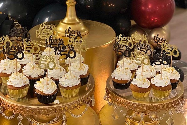 Cupcakes with white frosting and gold decorations displayed on gold stands, each topped with “cheers to 50 years” toppers, surrounded by black and gold balloons.