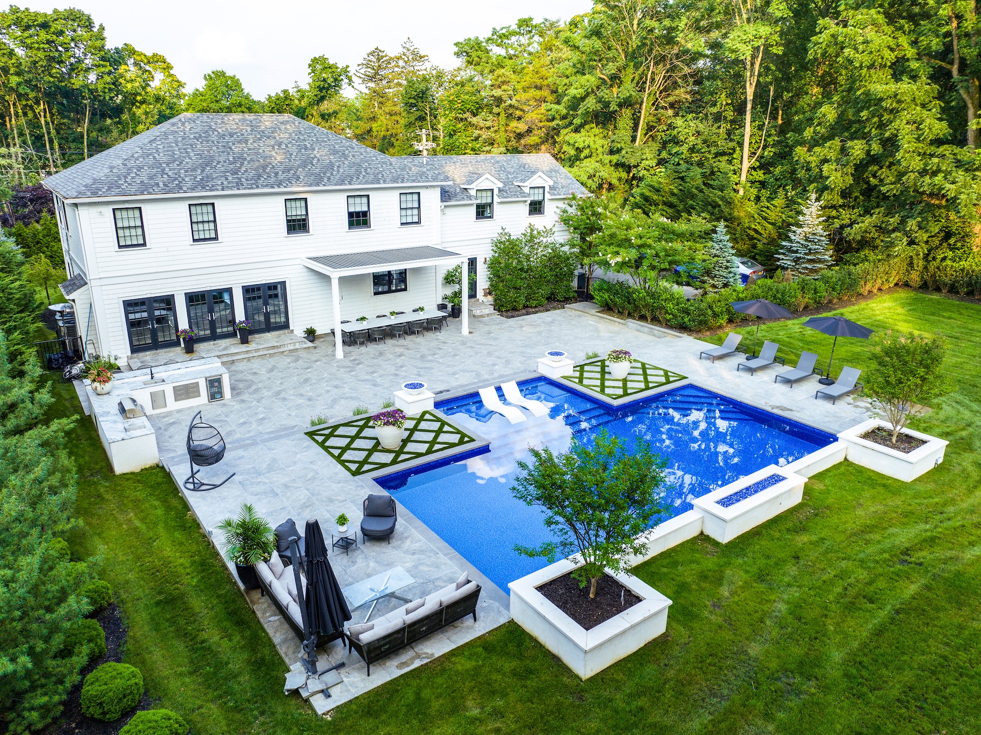 Aerial view of a large modern backyard with a rectangular pool, lounge chairs, patio seating, and landscaped greenery behind a two-story white house.