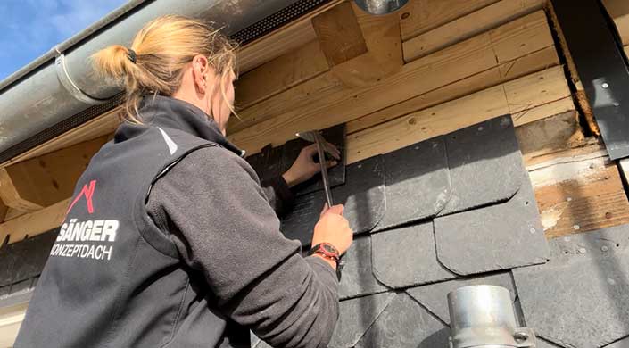 Eine Person befestigt mit einem Hammer schwarze Schieferplatten auf einem hölzernen Dach.