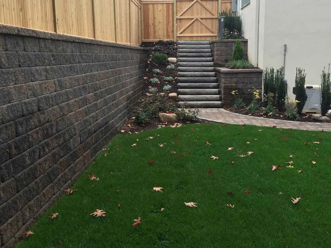 A landscaped backyard features a stone retaining wall, a patch of green grass with scattered fallen leaves, and stone stairs leading up to a wooden gate in Elwood.