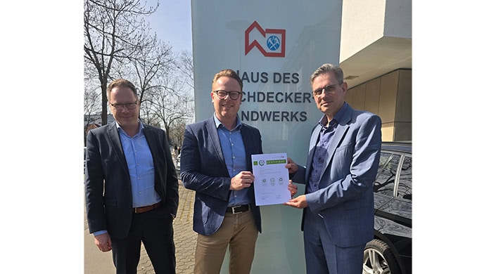 Ein Mann steht vor einem Gebäude und hält ein Zertifikat. Im Hintergrund ist ein Schild mit der Aufschrift "Haus des Dachdeckerhandwerks" zu sehen.