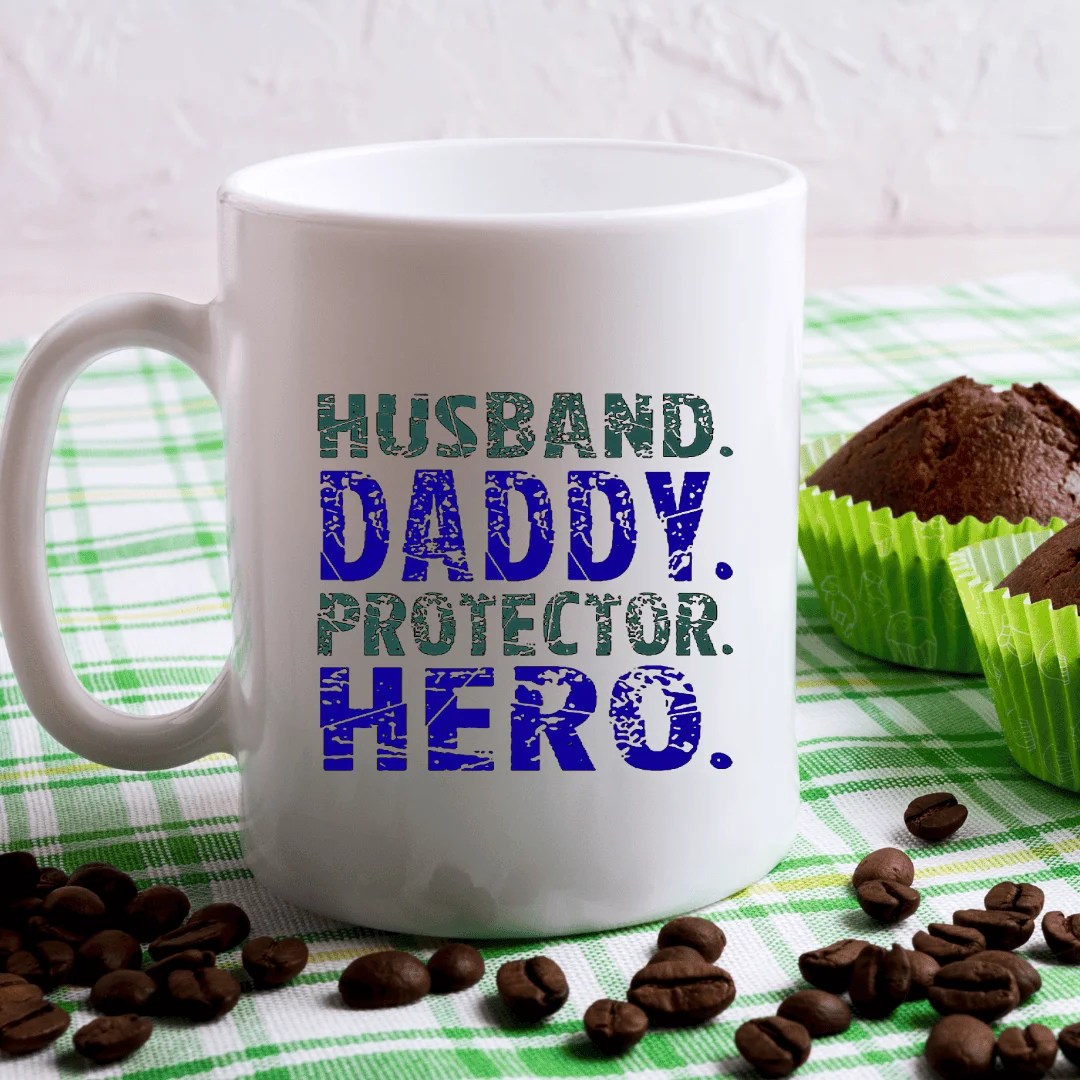 A white mug with bold blue and green text reading Husband Daddy Protector Hero sits on a green-checked cloth beside coffee beans and two chocolate muffins in green wrappers.