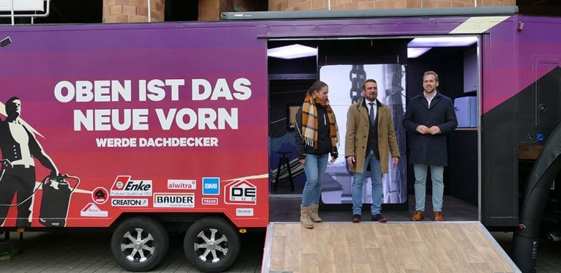 Eine Frau steht vor einem lila Wagen mit offenem Eingang, trägt eine karierte Schal und Winterkleidung.