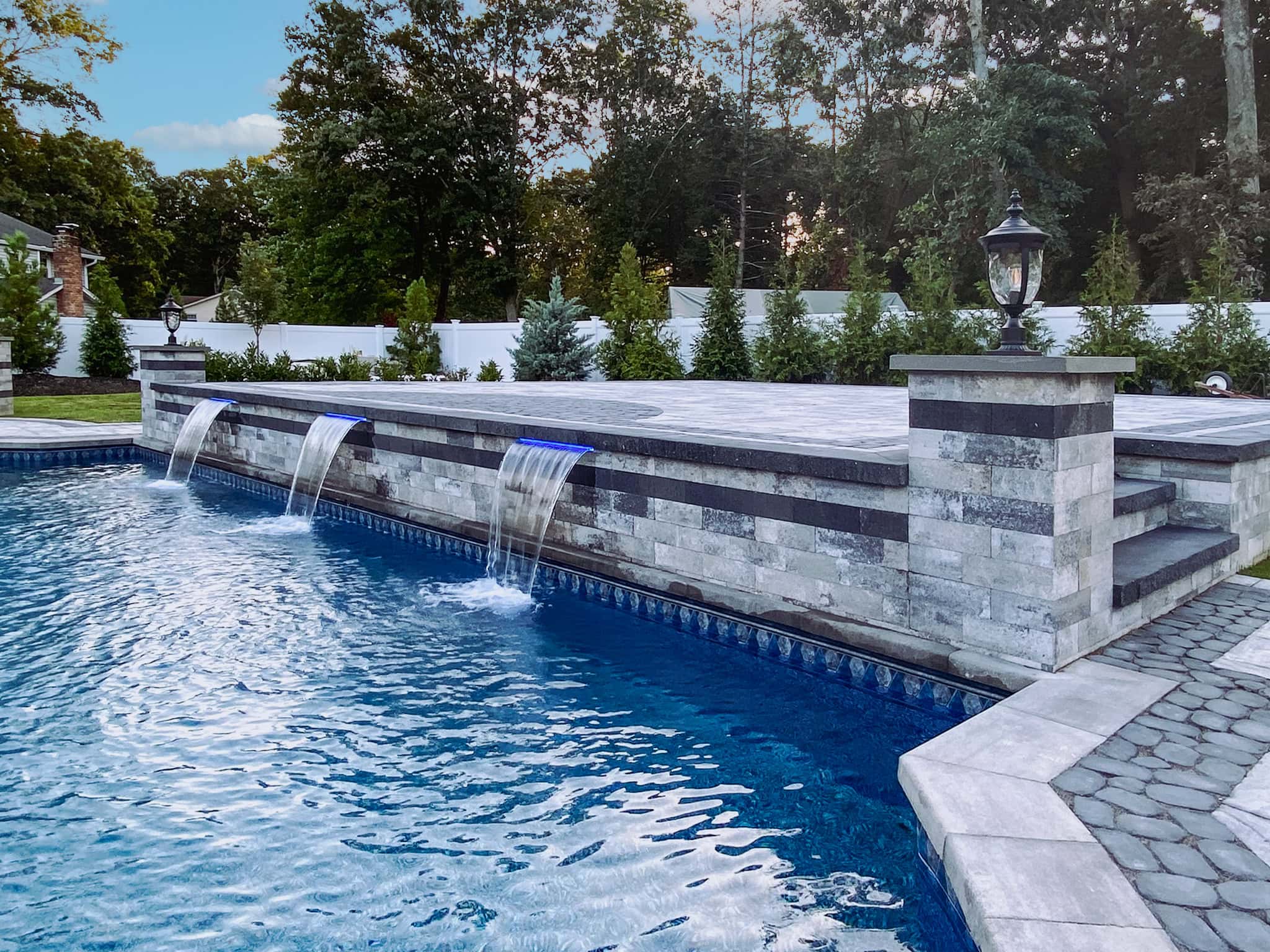 A modern rectangular swimming pool with water features flowing from a raised stone wall, surrounded by trees and a stone patio.