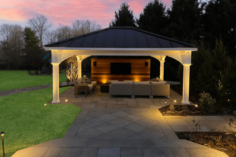 A covered outdoor patio with sofas, a TV mounted on a wooden wall, and accent lighting sits beneath elegant pergolas in a landscaped backyard at sunset.