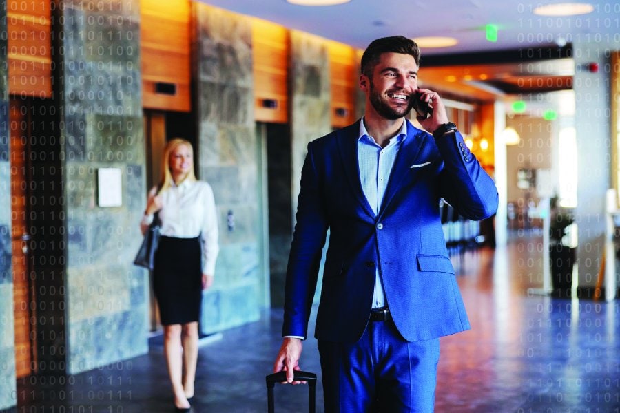 **Alt Text:** A professionally dressed man in a blue suit walks through a modern hotel lobby while talking on his smartphone. He pulls a small suitcase, suggesting business travel. In the background, a woman in business attire walks past elevator doors. Faint binary code overlays the image, symbolizing data, connectivity, and technology in the context of travel or hospitality.