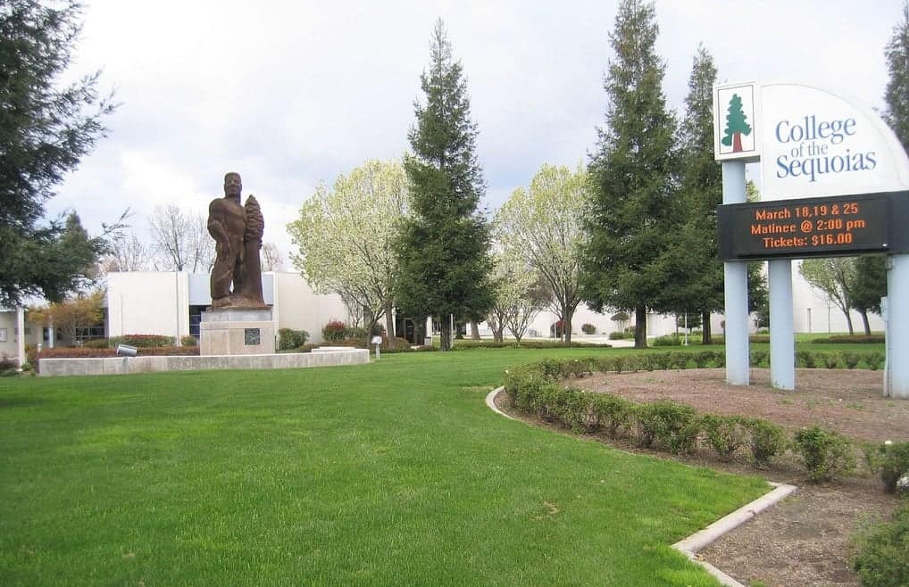 The entrance sign of College of the Sequoias on a green lawn in front of campus buildings and trees under a cloudy sky.