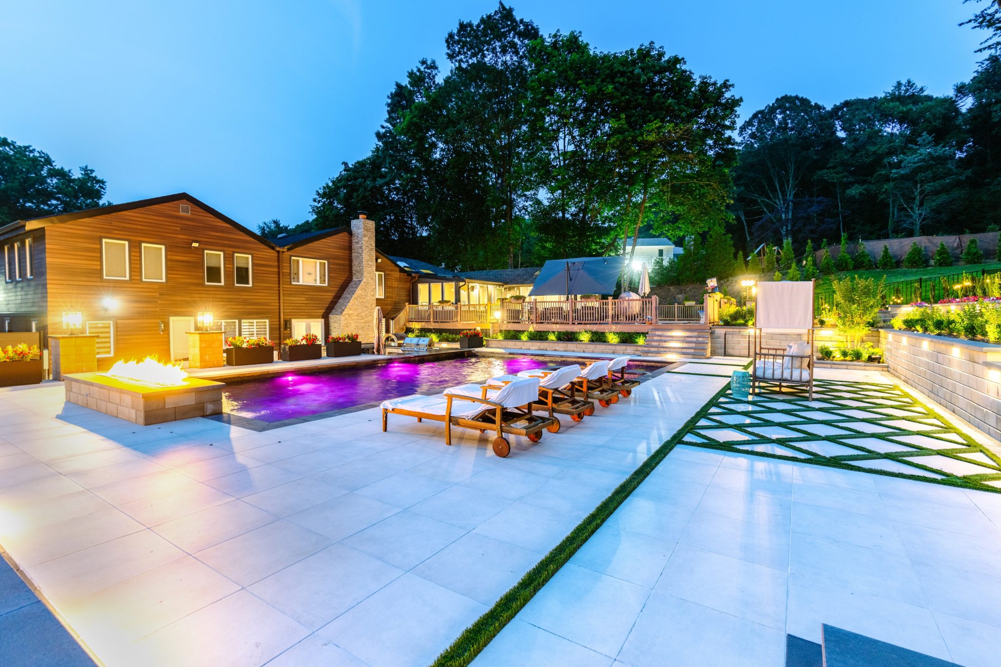 Modern backyard with a lit swimming pool, lounge chairs, fire pit, and landscaped patio area beside a large house at dusk.