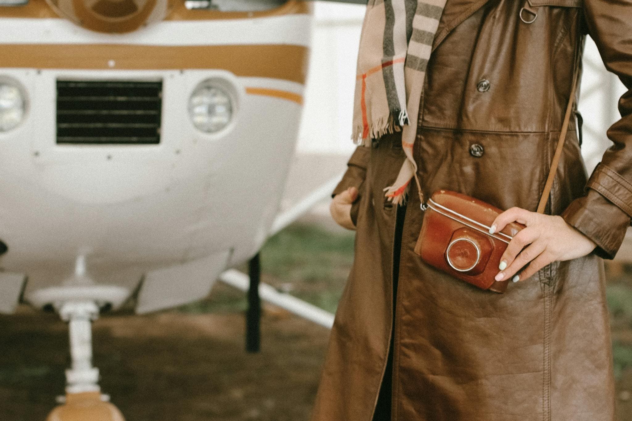 Stylish leather trench coat with cross body bag beside a small airplane outdoors.