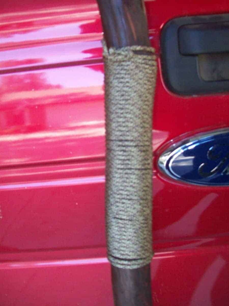 Close-up of a hand-wrapped wooden staff against a red background, possibly a car door. The staff, likely a handmade walking stick or oak staff for walking, is wrapped in a textured, light brown material. The wrapping technique appears to be hand done, showcasing wooden crafts techniques. The red background suggests a public park oak staff creation.