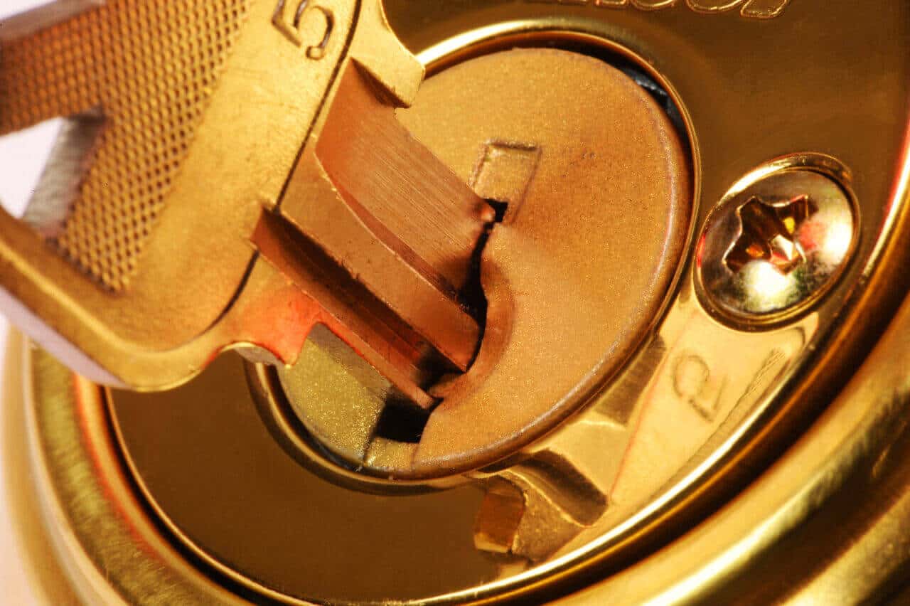 Close-up image of a key partially inserted into a brass lock. The key and lock both exhibit a metallic gold color, reflecting the craftsmanship of an expert locksmith. The lock is surrounded by a circular brass plate, and there is a screw visible to the right.