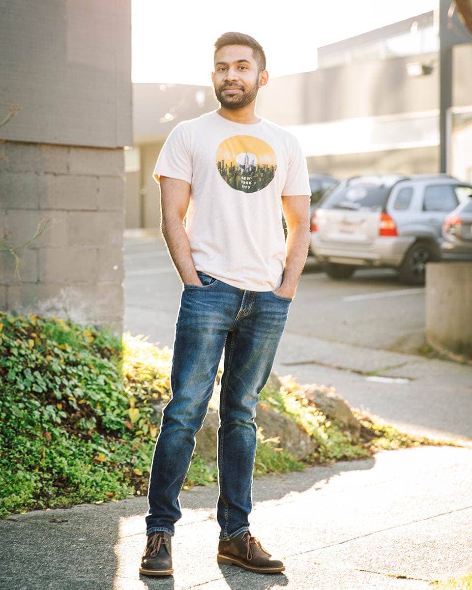 A man stands outdoors on a sidewalk with hands in his pockets, wearing jeans and a printed T-shirt; cars and buildings are visible in the background.