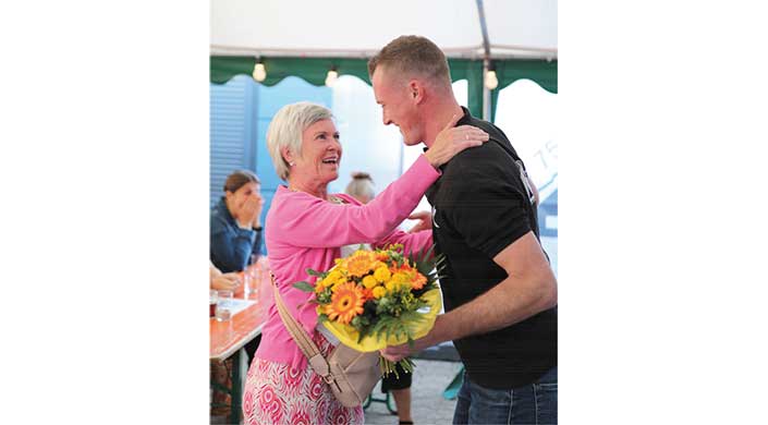 Eine ältere Frau in rosa Jacke umarmt einen jüngeren Mann und hält einen bunten Blumenstrauß. Beide lächeln sich an, im Hintergrund verschwommene Personen.