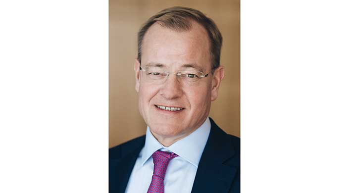 Ein Mann mit Brille, kurzem Haar und Anzug mit lila Krawatte lächelt in die Kamera.