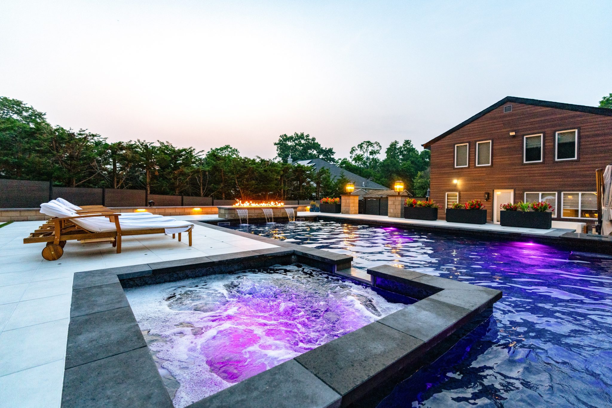 Modern backyard with a large pool, hot tub with purple lighting, wooden loungers, and a two-story house at dusk.