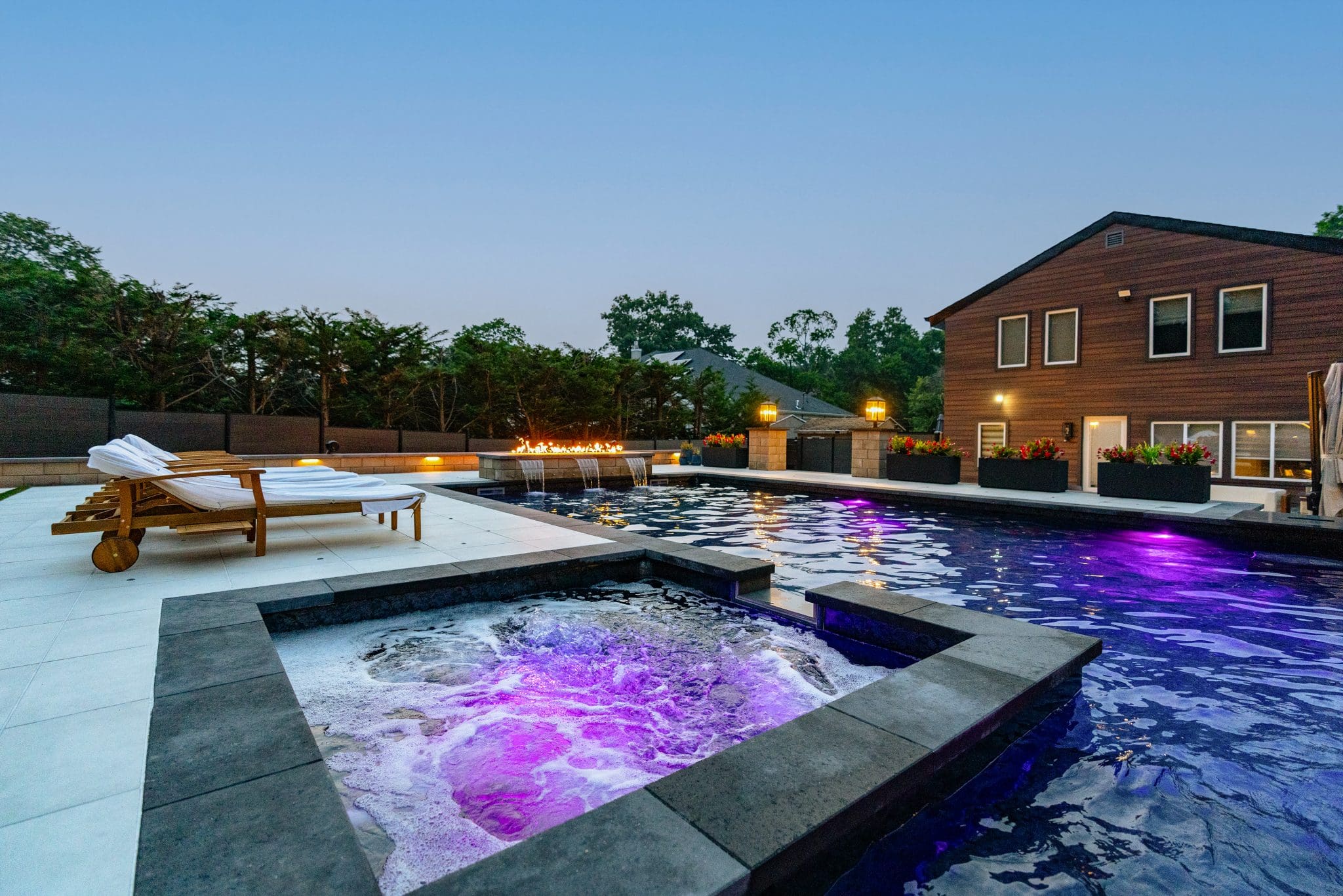 Modern backyard with a glowing hot tub, large swimming pool, lounge chairs, and a two-story house at dusk.