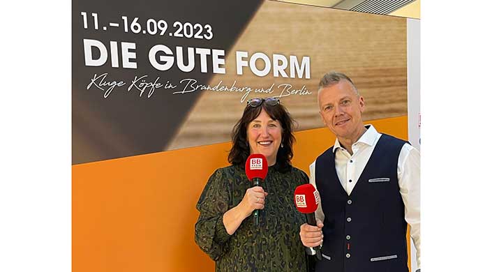Eine Frau mit dunklen Haaren hält ein rotes Mikrofon, steht lächelnd vor einem Banner mit der Aufschrift „GUTE FORM 2023“ und trägt ein grünes gemustertes Oberteil.