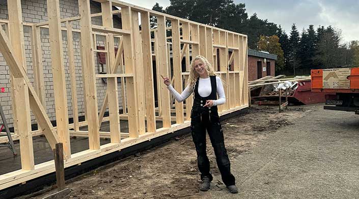 Eine Frau steht auf einer Baustelle und zeigt auf ein Holzrahmengerüst eines im Bau befindlichen Gebäudes.
