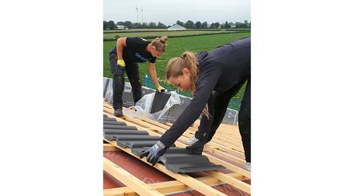 Zwei Frauen legen Dachziegel auf einem Hausdach, arbeiten konzentriert und tragen Handschuhe. Im Hintergrund ist ein Feld und eine Scheune zu sehen.
