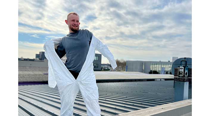 Person in weißem Schutzanzug steht auf einem Wellblechdach, im Hintergrund Gebäude und bewölkter Himmel.