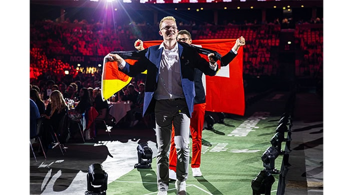 Ein junger Mann in Anzugjacke und grauer Hose steht auf einer Bühne und hält eine große rot-weiße Flagge.