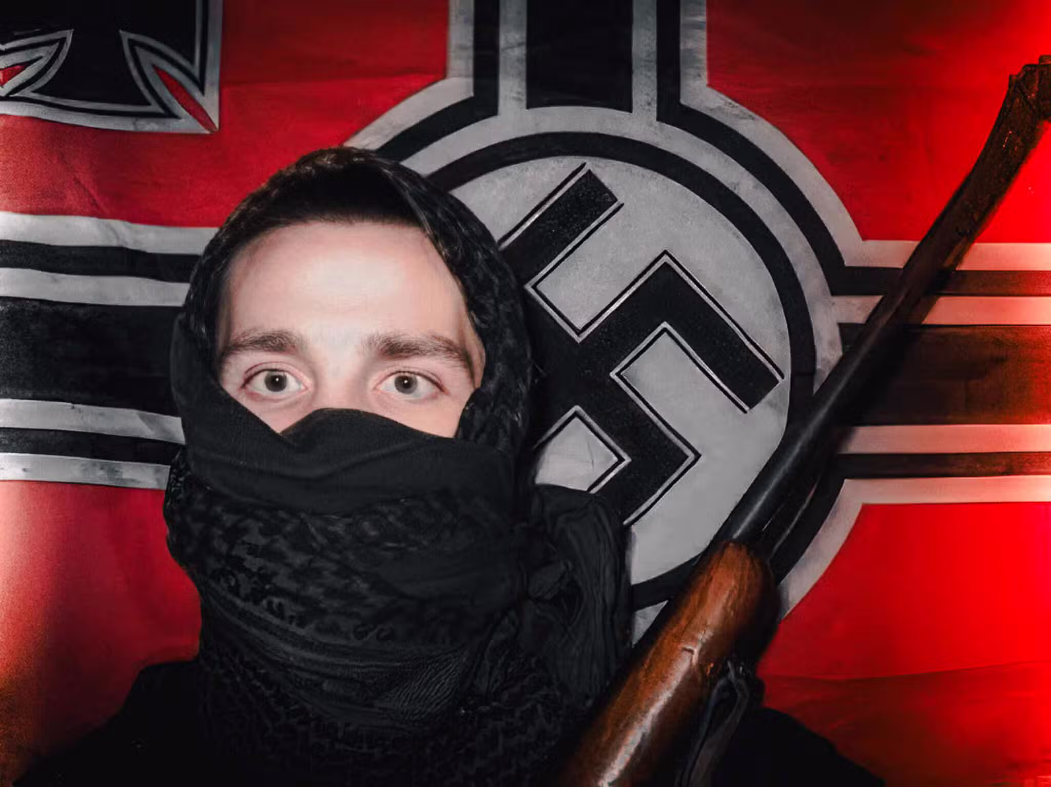 A man with his face covered by a black scarf stands in front of a Nazi-era war flag.