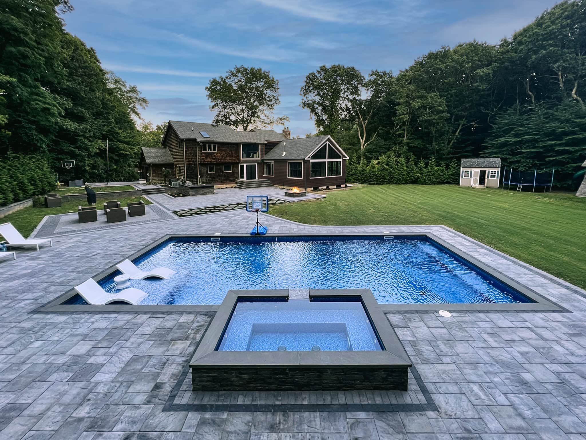 A rectangular in-ground pool with a built-in hot tub is surrounded by patio seating in the backyard of a large house with a lawn, trees, and a shed.