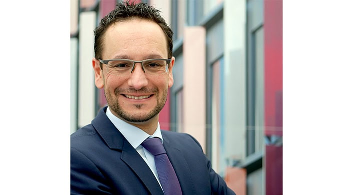 Ein Mann im Anzug und mit Brille lächelt, im Hintergrund sind unscharfe Bürogebäude zu sehen.