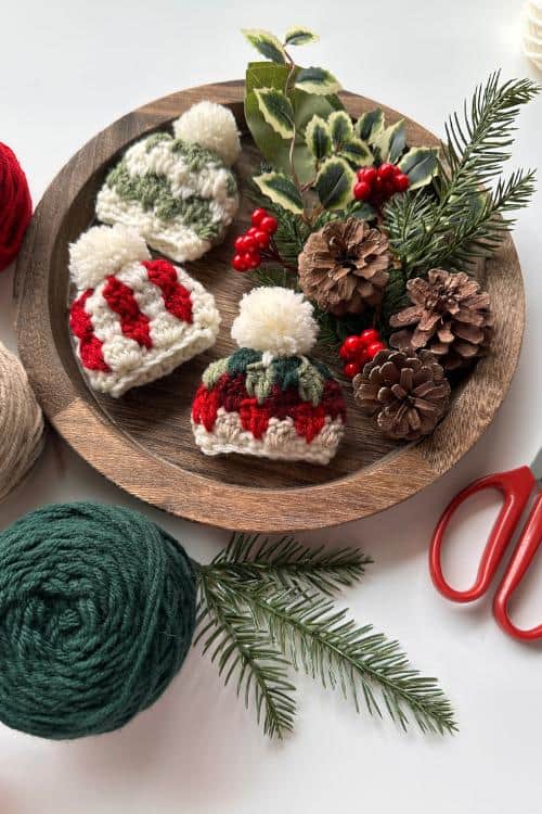 A wooden plate holds three small wool hats, pinecones, holly, and red berries amid yarn balls, pine branches, and red scissors.Free crochet mini hat christmas ornament pattern