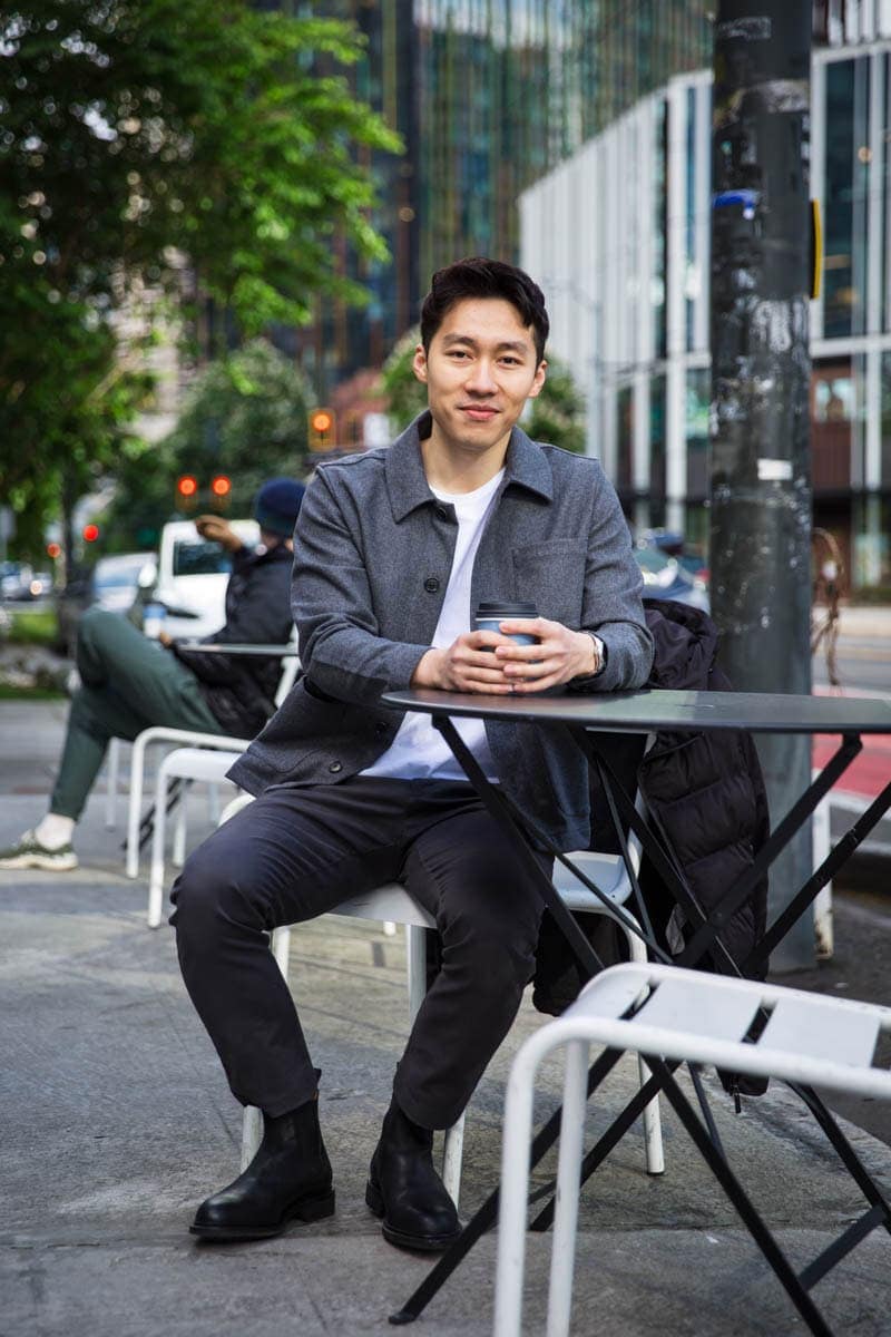 Man sitting outdoors at a café table, holding a coffee cup, with a street and buildings in the background.