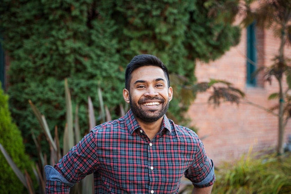 A man with a beard wearing a red and blue plaid shirt stands outdoors, smiling in front of greenery and a brick wall.
