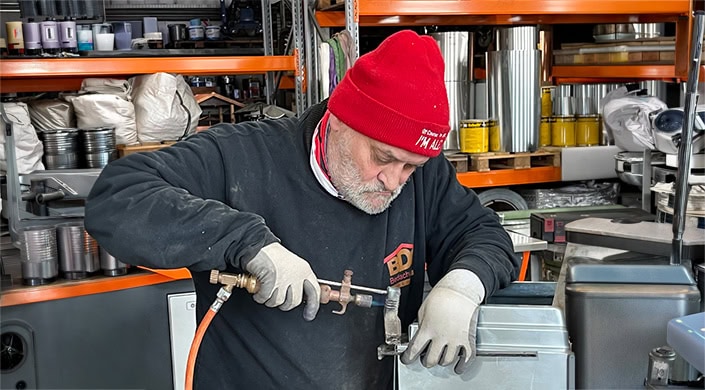 Ein älterer Mann mit roter Mütze arbeitet konzentriert mit einem Brenner an einem Metallteil in einer Werkstatt.