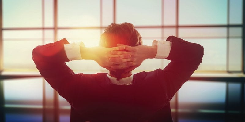 A man in a business suit sits relaxed with his hands clasped behind his head, gazing through a large window that reveals a warm, glowing sunset. The lighting creates a serene and contemplative atmosphere, emphasizing a moment of relaxation and reflection. The blurred indoor setting contrasts with the vibrant outdoor view, symbolizing a pause amidst a busy professional life.